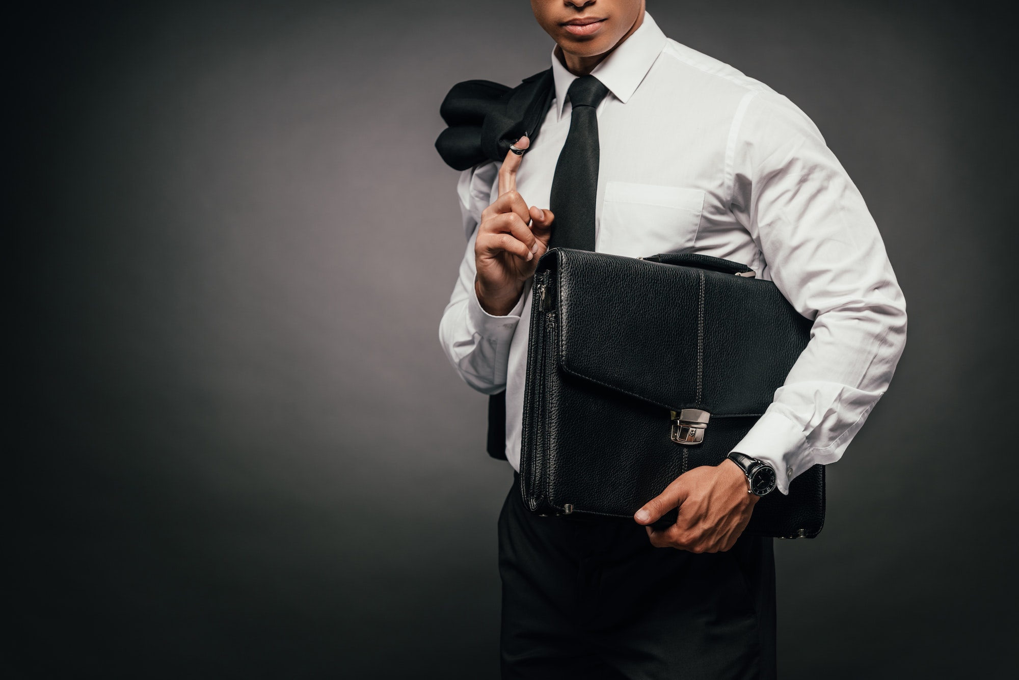 cropped view of african american businessman holding blazer and leather briefcase on dark background